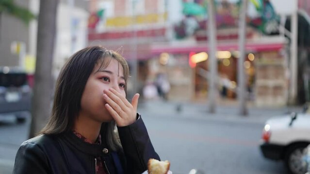 A young Japanese woman from Okinawa Prefecture in her 20s in winter clothes eating street food lots of sataandagi on Kokusai Street in Naha City, Okinawa Prefecture 沖縄県那覇市の国際通りでストリートフードのサーターアンダギーを食べる冬