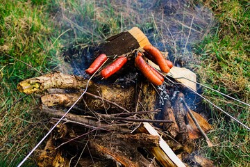 Traditional-style roasted sausages over a grassy bonfire with wood embers, emitting white smoke. Sausages exhibit a beautiful golden-brown color.