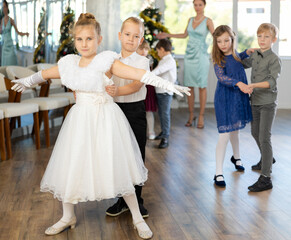 Enthusiastic preteen children, girls and boys in elegant outfits practicing partner dance moves in studio adorned with Christmas decorations, under guidance of female teacher