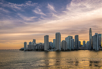 Miami, Florida, USA - July 29, 2023: Wide shot. Sunset sky over buildings on Brickell Key island at evening 19:43. Tequesta points in center. Both sides of river mouth.. Yellow water