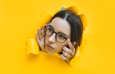Portrait of a cute young woman with glasses looking through a hole in yellow paper. An incredulous...