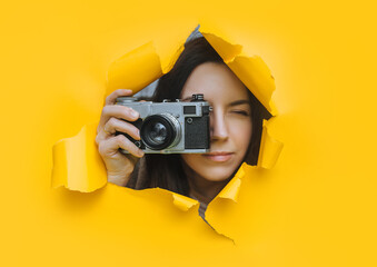 A young paparazzi girl with a rare SLR camera looks out from her hiding place and carefully watches what is happening. Yellow paper hole.Tabloid press.Looking for a subject for stock photos - obrazy, fototapety, plakaty