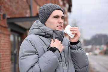 Sick young man with tissue on city street. Cold symptoms