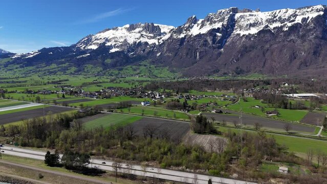 Aerial establishing shot of highway in rural village of Liechtenstein. Beautiful idyllic fields and snowy alps in background. Sunny day on border between Switzerland and Lichtenstein.