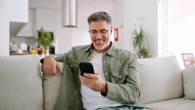 Smiling happy senior mature middle aged man holding cell mobile phone using smartphone sitting at home on couch, scrolling social media, checking financial apps, buying online, texting messages.