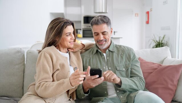 Happy older mature senior family couple, middle aged man and woman looking at cell phone using smartphone mobile technology device together sitting at home relaxing on couch doing online shopping.
