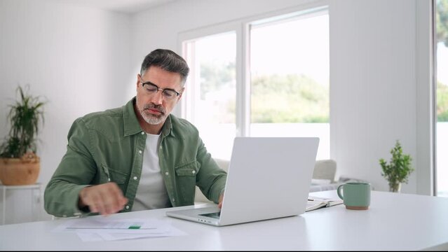 Happy middle aged mature man entrepreneur wearing eyeglasses looking at laptop holding paper bill checking financial invoice or tax document making online payments working at home.