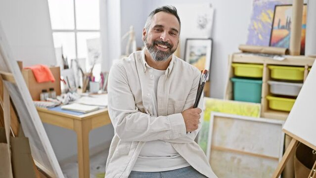 A smiling mature man with a beard and grey hair holding paintbrushes in an art studio interior.