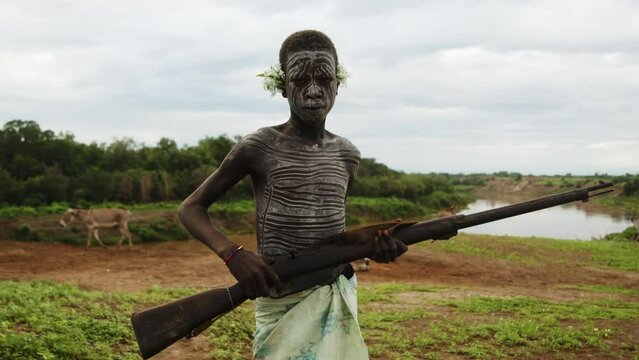 Kid with Face Paint from Karo Tribe with a flower on his ear holding a rifle