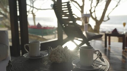 Static shot 2 white steaming mugs on a small table in a tropical luxury villa with the sunny ocean in the distant background.