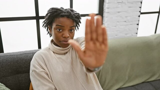 Unyielding young black woman with dreadlocks, perched on home sofa, stoutly gesturing 'stop' with firm palm. her face a mask of serious denial, a warning of her refusal to accept the negative.