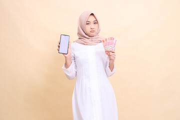 Young Asian Muslim woman wearing a hijab frowns holding a cell phone gadget and rupiah money in her hand. Lifestyle, business and commercial concept