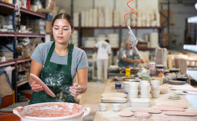Young woman ceramist painting crafted plates by dipping it into bucket of color.