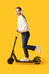 Happy young Asian woman in headphones riding modern electric kick scooter on yellow background