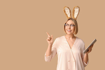Happy adult businesswoman in bunny ears with tablet computer pointing at something on beige...
