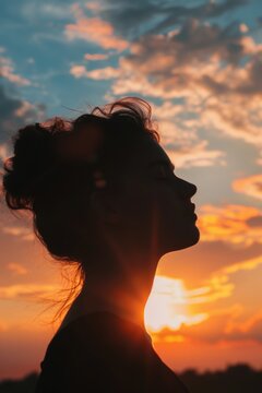 A woman is standing in the sun with her back to the camera. The sun is setting in the background, casting a warm glow on her face. Concept of peace and tranquility