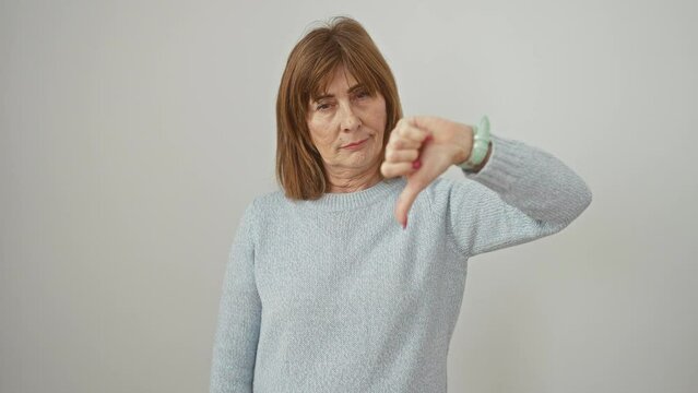Angry looking middle age woman wearing sweater gives a bad, negative thumbs down gesture, expressing unhappy rejection on isolated white background.