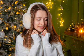 Close-up portrait of a young woman dressed in a white sweater and warm headphones posing against a background of golden bokeh and a Christmas tree.