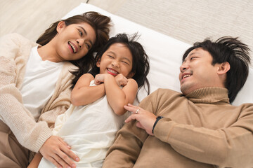 Happy Asian family, father, mother, cute daughter having fun, lying on bed in bedroom at home, bond and relax together, smiling with happiness. Top view