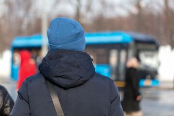 Back view of a man in a blue hat and black jacket waiting for a bus