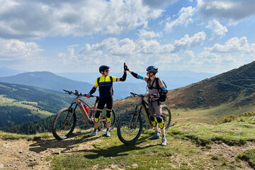 Two cyclists men riding electric bikes outdoors. Male tourists resting on the top of hill, giving...