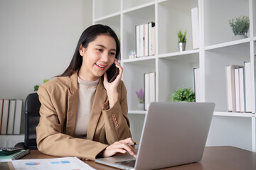 Fototapeta premium Young Asian business woman sits on the phone in an online business meeting using a laptop in a modern home office decorated with shady green plants.