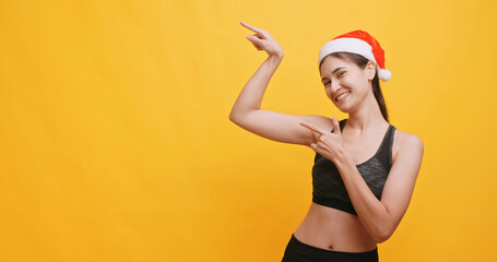 Young sportswoman wearing a Santa hat shows physical strength and points at something. Isolated on...