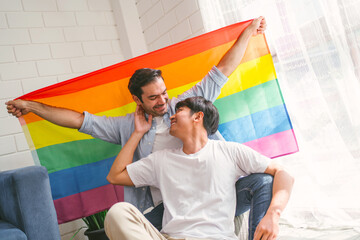 Happy caucasian and Asian LGBT couple, sitting on the sofa holding and waving rainbow LGBT Pride flag together in the living room at home. Diversity of LGBT relationships. A gay couple concept.