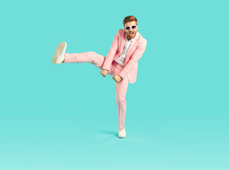 Full body studio portrait of a funny dancing man. Full length happy, cheerful young party lover wearing a trendy pink suit and sunglasses dancing isolated on a bright turquoise blue colour background
