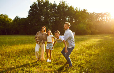 Naklejka na ściany i meble Happy young family of four having fun and playing plane at sunny day in nature. Mother, father and their children boy and girl walking in the park on green grass outdoors at sunset together.