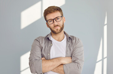 Portrait of confident serious bearded handsome man in glasses standing with crossed arms wearing white t-shirt and beige shirt isolated on grey wall background. Smart guy looking at the camera.