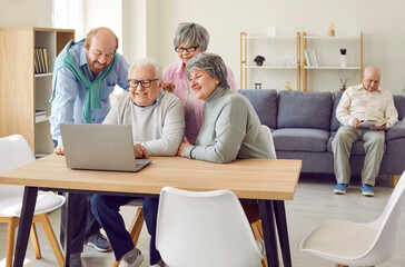 Obraz premium Group of elderly people use laptop to watch movies, read news and make video calls to relatives. Cheerful senior male and female pensioners sitting at table in front of laptop in bright living room.
