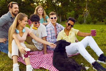 Happy friends having fun playing with dog while sitting on green lawn in park. Group of multiracial male and female best friends enjoying picnic outdoors. Friendship, leisure activities concept