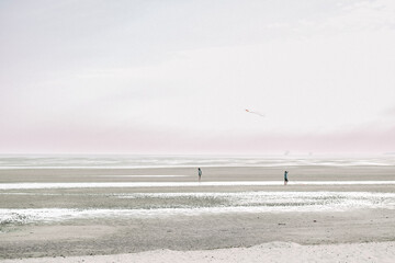 People fly a kite on the seashore on a cloudy day.Wadden Sea Coast.Frisian Islands.Fer Island.Germany.Kites and people walking on the white sandy sea beach.Flying kites on the seashore. - obrazy, fototapety, plakaty