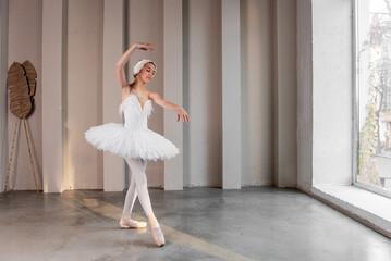 Elegant young ballerina performs beside large windows, poised stance, delicate white tutu illuminated by natural light, embodying the fusion of strength, grace. White swan performance. Copy space