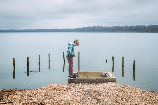 Boy looking into spillway on shore of lake on cloudy winter day