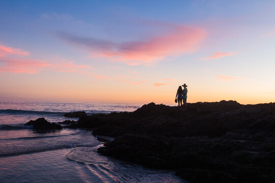 Two people stand on rocks at the beach at sunset.