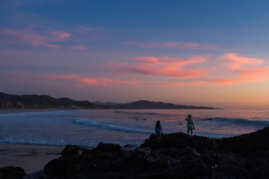 Two people stand on rocks at the beach at sunset.