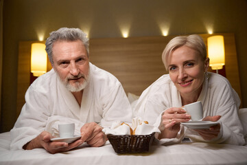 Front view of adult couple drinking tea or coffee and looking at camera on bed