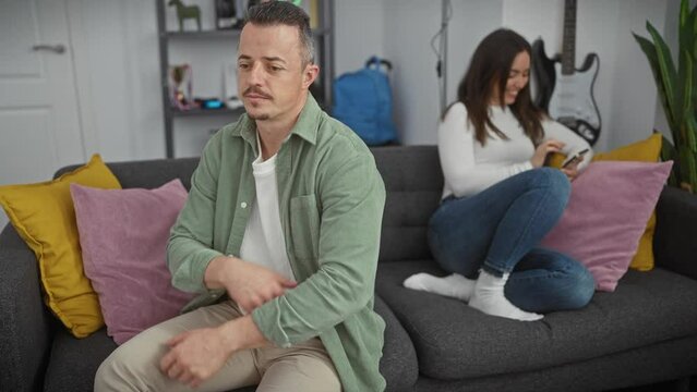 Worried man sitting, woman using smartphone in living room.