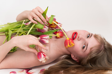 Obraz na płótnie Canvas Playful half naked woman is posing with bouquet of spring flowers tulips for Woman's Day on her chest and in hands. Sensuality gen z woman looking at camera and holding flowers, lying down on white