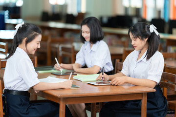 Three Asian female high school students sat together reading books and looking at laptops on study tables together to study in preparation for exams in the library of a famous school.