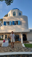 A man in casual attire stands confidently in front of a majestic white building, the sunlight...