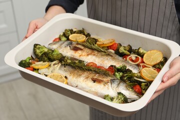 Woman holding baking dish with delicious fish and vegetables indoors, closeup