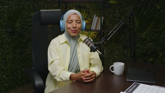 Portrait of young female podcast host in hijab and headphones sitting with teacup at desk with microphone, looking at camera and smiling in recording studio
