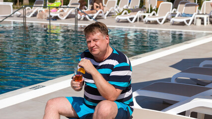 A plump man of European appearance with an alcoholic drink sits by the pool. - obrazy, fototapety, plakaty