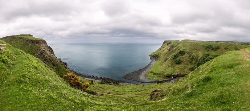 The coastline near Lealt Falls on the Isle of Skye is captured in a panoramic view, featuring lush hills that gracefully descend to a pebble-strewn beach and the calm sea beyond