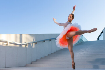 One Graceful Slim Professional Caucasian Ballet Dancer in Rose Pink Tutu Dreass Whie Posing In Dance Flying Pose On Blue Stairs In Stretched Pose Outdoor. - 770484545