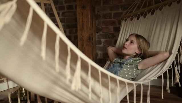 Preteen girl child lying in hammock and singing song at home. Pretty fe,ale kid relaxing in loft room