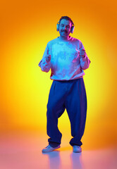 Positive, energetic, smiling man in pink sweatshirt and jeans listening to music in headphone and dancing on gradient yellows orange background in neon. Concept of human emotions, casual fashion